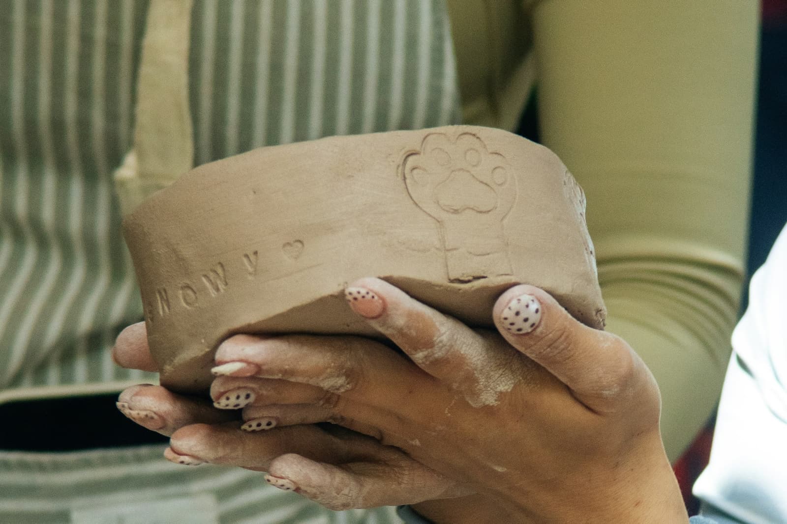 Clay-dusted hands holding an unfired clay bowl, stamped by hand with a paw print and a pet’s name